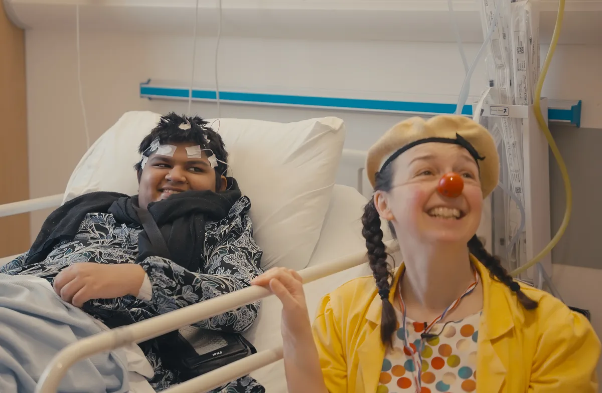 In a hospital room a young person sits up in bed smiling alongside Dr Wallop