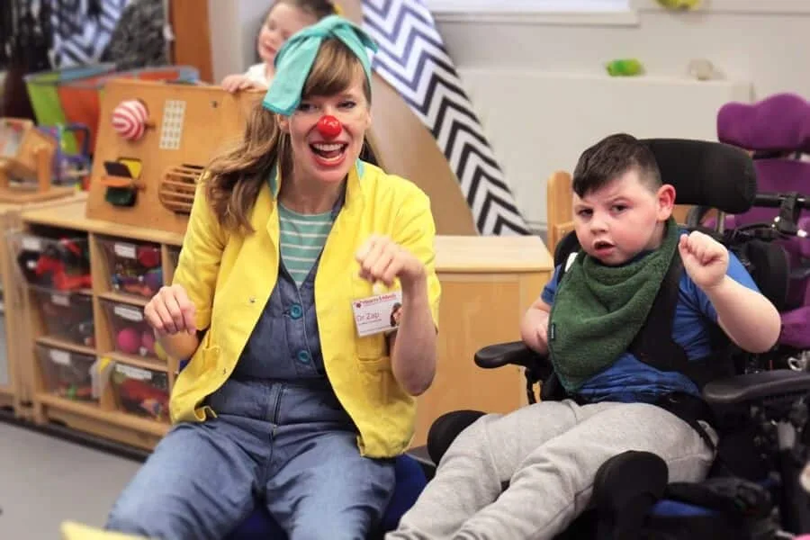 Dr Zap and a young person are sitting on a chair and in a wheelchair and are looking at something. Their hands are closed in fists like riding a bike