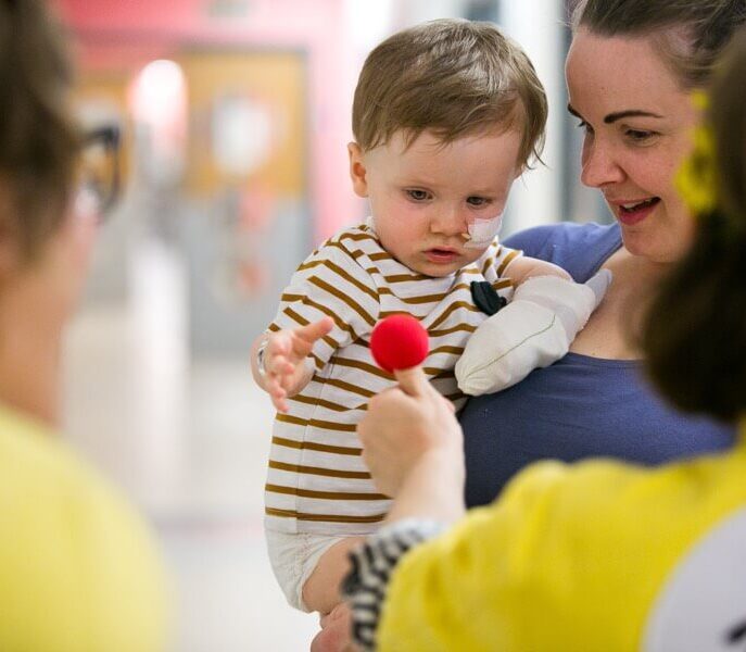 In hospital. A child with a nasogastric tube is held by a woman and is about to grab a sponge red nose from a Clowndoctors thumb
