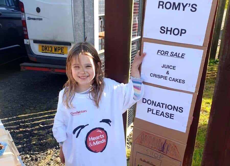 A child in an over-sized Hearts & Minds t-shirt stands next to a sign for Romy's Shop. Offering juice & Crispie cakes for donations