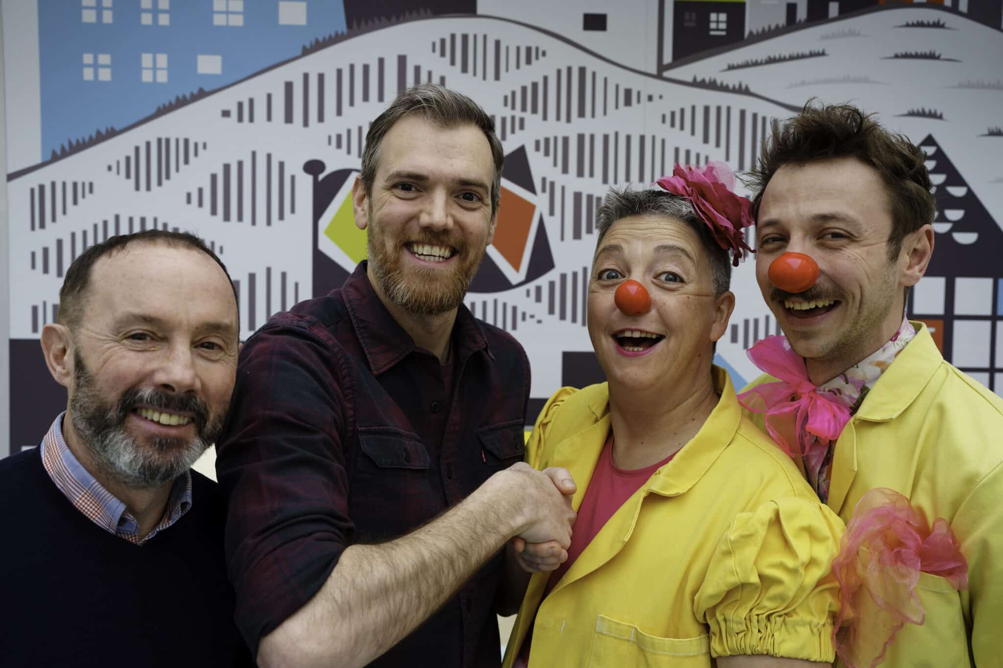 4 people are standing side by side and smiling to the camera. the two men on the left are wearing casual clothes and the man and woman on the right are wearing yellow doctor coats bright pink t-shirts and scarves and red clown noses. They are both Clowndoctors. The man and the lady Clowndoctor in the middle are shaking hands.